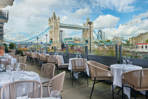 A group of tables and chairs outside a restaurant overlooking London Bridge and the Thames river.